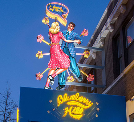 A neon sign at dusk featuring a man and a woman in 1950s-style clothes dancing. Stars surround them and a word bubble says "I found my thrill" and below in yellow script it says "Blueberry Hill", the name of the iconic St. Louis restaurant and music club