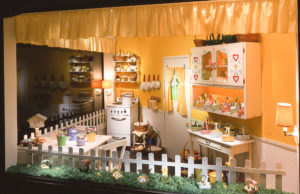 A large corner display window that feature's the Easter Bunny's kitchen. There are yellow walls, old-fashioned white stove, cabinets, and table, and there are buckets of colorful Easter eggs in colorful pails on the table. There are boiling eggs on the stove. There is a white picket fence on the ledge with green paper grass and Easter eggs.