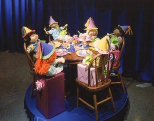 Large corner display window featuring cabbage patch kids of different races having birthday party, sitting around a small table with cake, birthday colorful hats, and noisemakers.