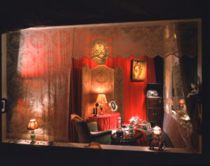 Photo of an elaborate corner display window which features a Victorian era apartment at Valentines Day. There are old lamps, an old chaise with a silk slip, chocolates, and Victorian furniture. There is lace draped everywhere.