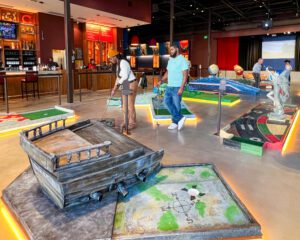 A woman and a man playing indoor miniature golf. The hole features a hand-built wooden pirate's ship.