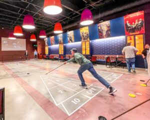 A man in green shirt and jeans playing indoor shuffleboard. Colorful magician-related posters are on the wall along with glowing display cases of golf-related memorabilia