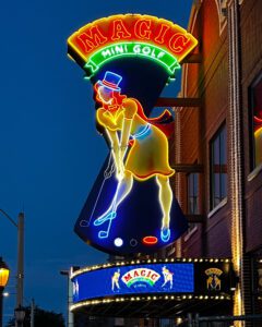 Huge 27' Magic Mini Golf neon sign at dusk. Big red neon circus style letters on a curved yellow neon-outlined background with a woman in yellow neon putting mini golf. She is wearing a yellow dress, a light blue magician's hat, and a red cape.