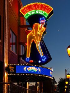 Huge 27' Magic Mini Golf neon sign at dusk. Big red neon circus style letters on a curved yellow neon-outlined background with a man putting mini golf. He is wearing a yellow tuxedo, a light blue magician's hat, and a red cape, all of which are outlined in neon.